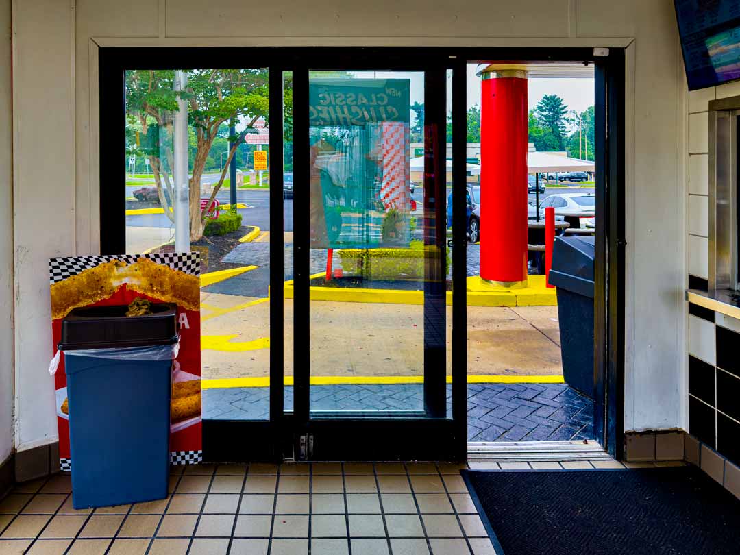 Fast-food restaurant lobby looking out the windows at the yellow curbs, the scarlet columns and posts, the red checkered banners and the green trees in the distance beyond the asphalt parking lot.